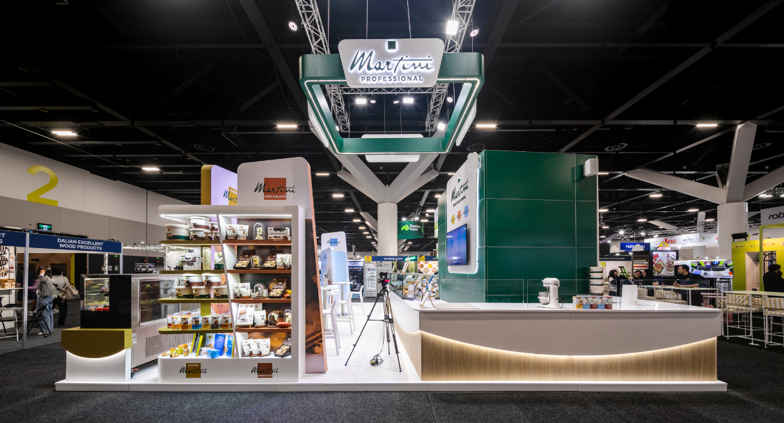 Custom island exhibition stand panorama with bespoke freestanding chocolate product shelving unit, full-height green feature wall, and curved demonstration counter built for Martini Professional at Restaurant & Foodservice Show Australia 2026