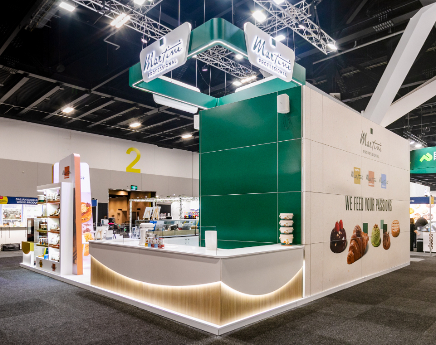 Custom fabricated exhibition stand corner with suspended hanging sign, full-height graphic wall panel, and curved illuminated timber-base counter built for Martini Professional at Restaurant & Foodservice Show Australia 2026